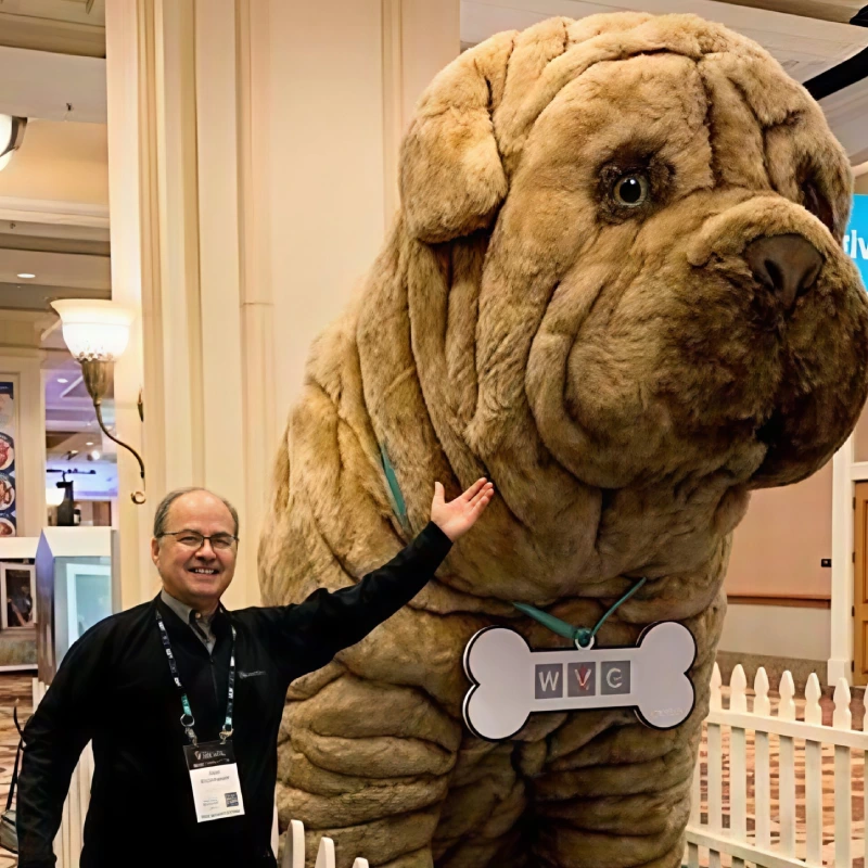 A man stands next to a giant plush dog that resembles a Shar-Pei, which is wearing a bone-shaped tag with "WVC" on it.