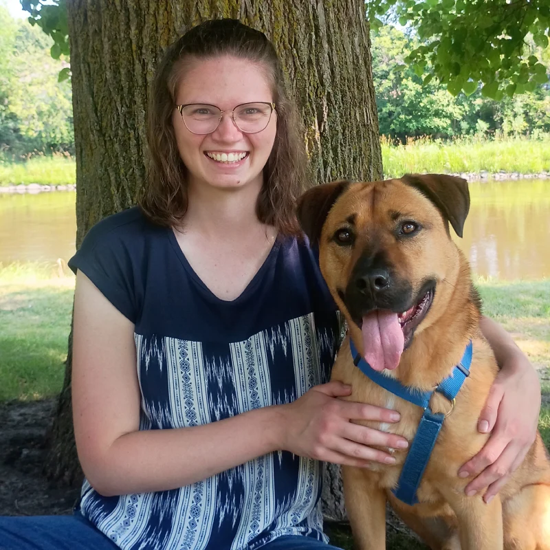 A person wearing glasses and a blue and white patterned shirt sits next to a large brown dog with a blue harness by a tree near a body of water.