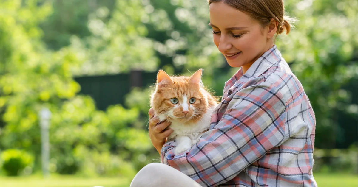 A person wearing a plaid shirt holds a fluffy orange and white cat in their arms while sitting outdoors on a sunny day.