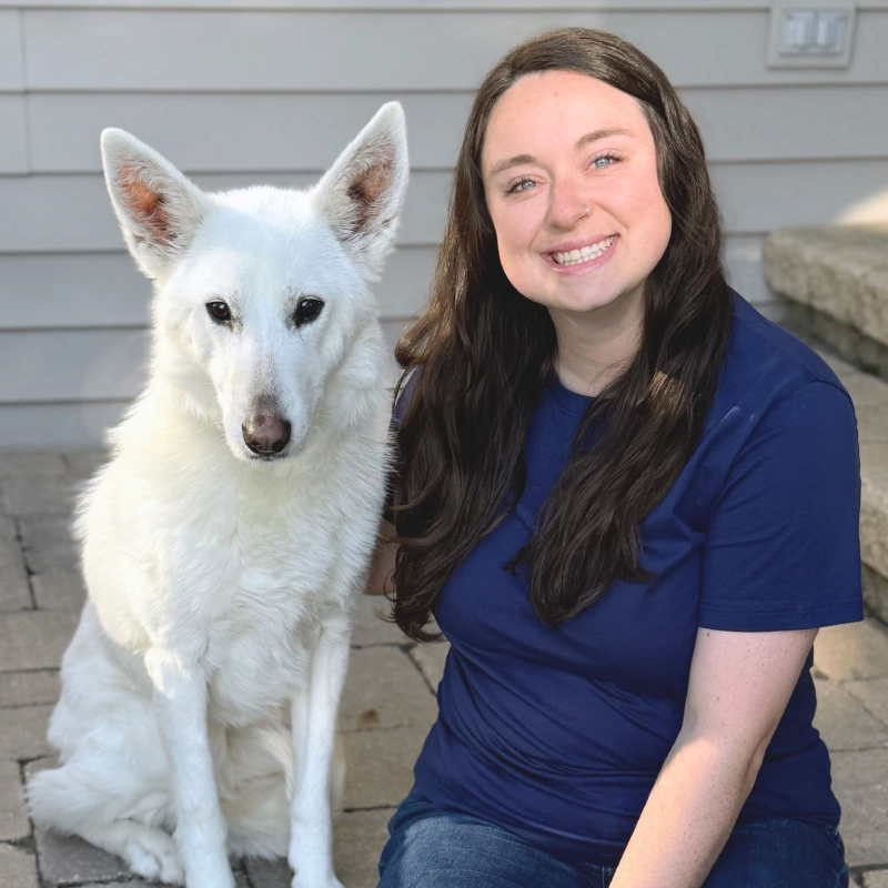 A person in blue scrubs kneels outdoors beside a white dog with upright ears, both facing the camera; the person smiles warmly while the dog sits calmly on the pavement.